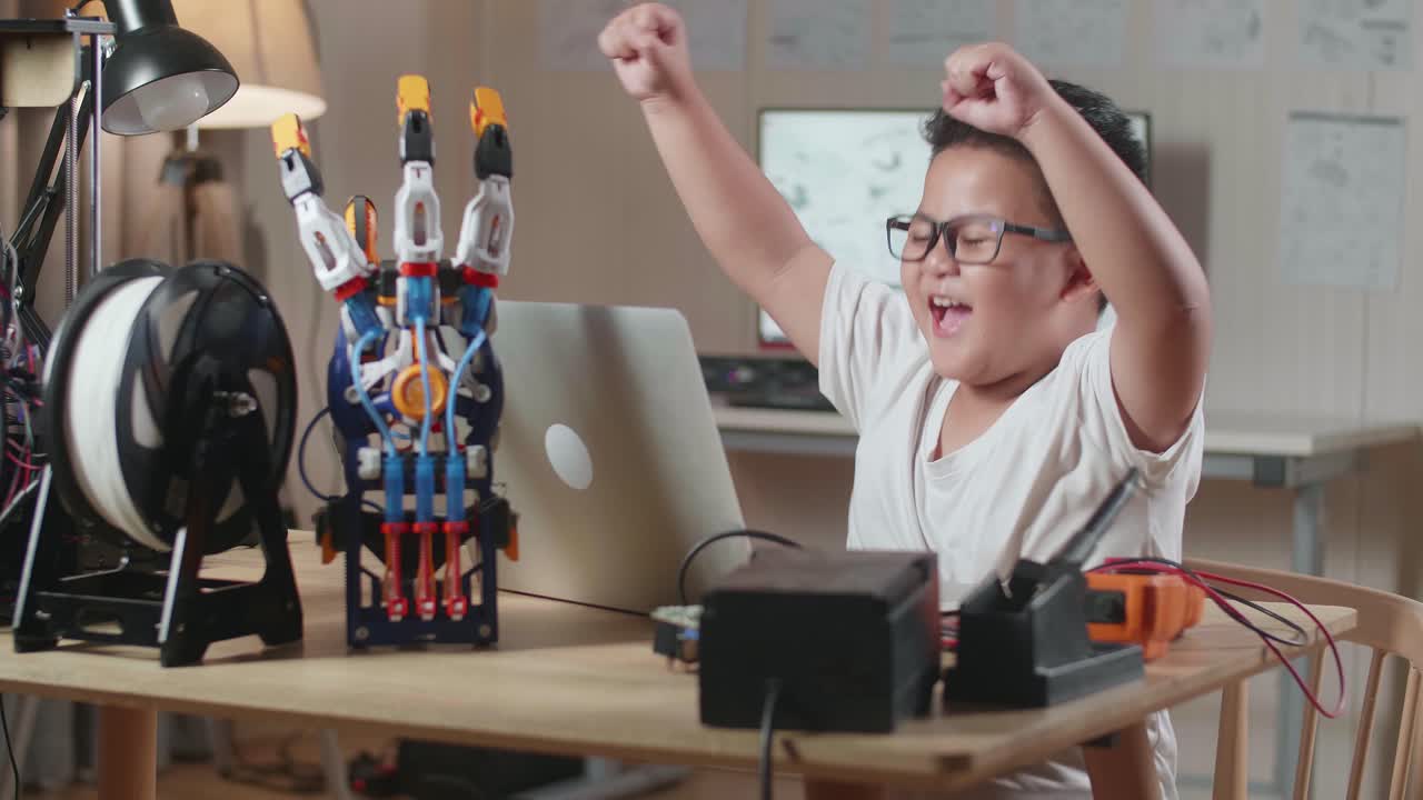 un chico asiático lindo celebrando el éxito trabajando sobre una mano cyborg en una computadora portátil en casa