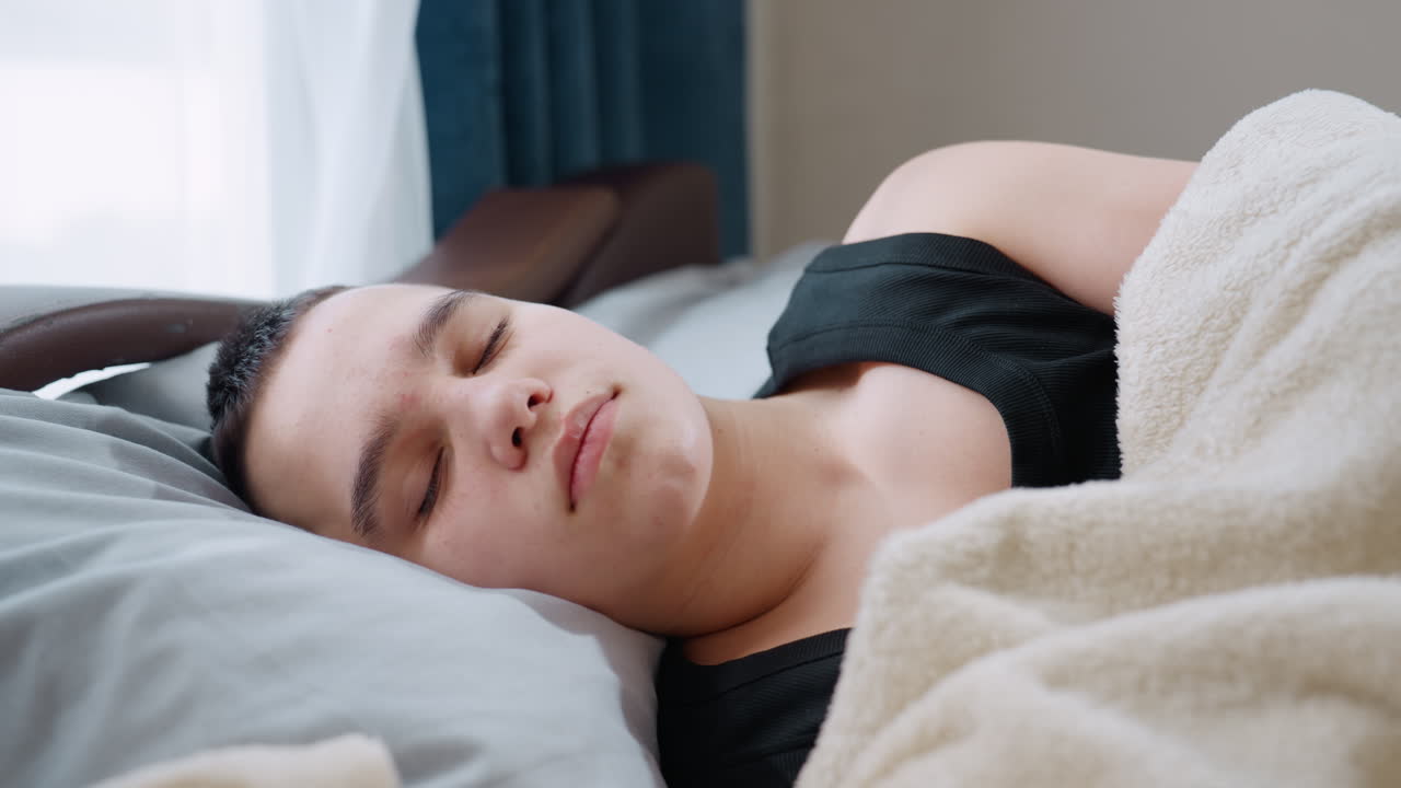 Peaceful boy rests under blanket lying on pillow with eyes closed enjoying calm sleep in bright cozy room with natural light from window highlighting comfort