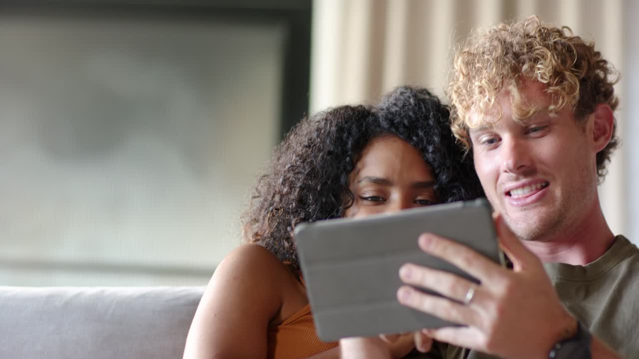Diverse couple leaning together on couch, sharing tablet folio held by man, laughing, reacting