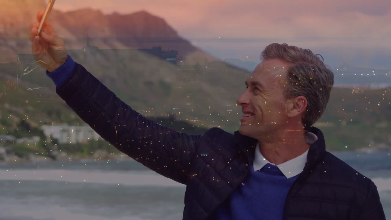 Video verticale: Uomo che fa un selfie con il telefono alzato in una baia al tramonto, con scintille animate sul telefono per un effetto tecnologico