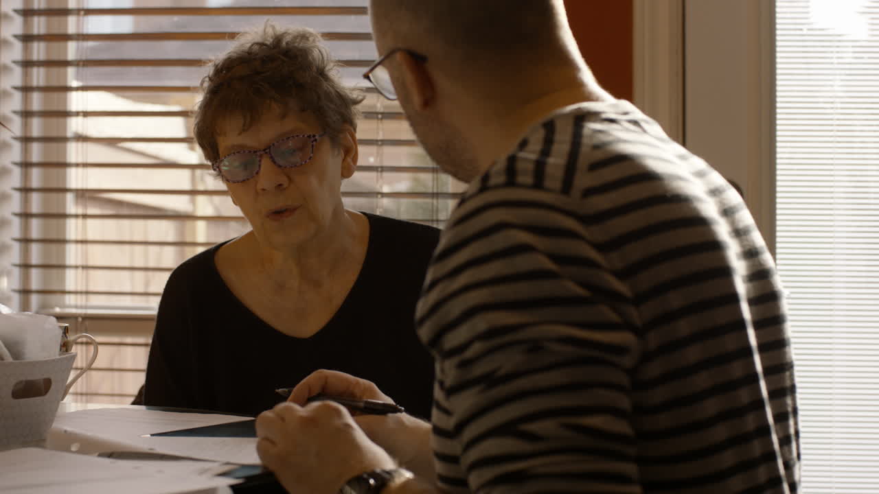 An elderly woman sits at a kitchen table with a middle-aged man, in a warmly sunlit house . He helps her fill out a paper questionnaire. She answers negatively, he writes with pen. Both wear glasses.