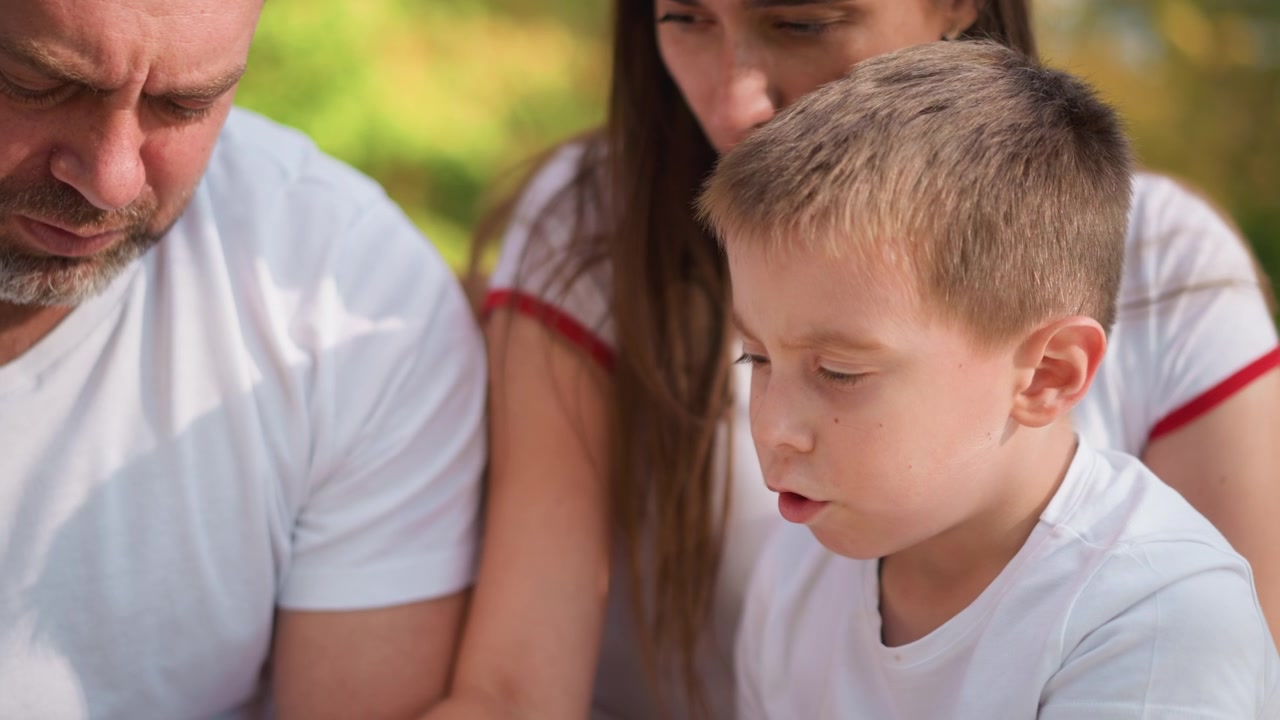 Primo piano di uomo, donna e bambino seduti insieme all'aperto in una luce soffusa. La famiglia indossa camicie bianche con bordo rosso su uno sfondo sfocato di fogliame verde. Interazione delicata.