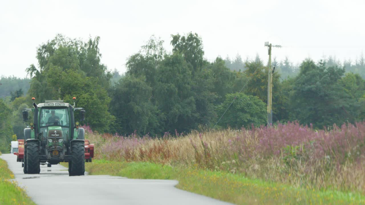 A green tractor travels along a narrow country road bordered by wildflowers and grass under overcast daylight, with steady frontal camera movement and natural rural scenery