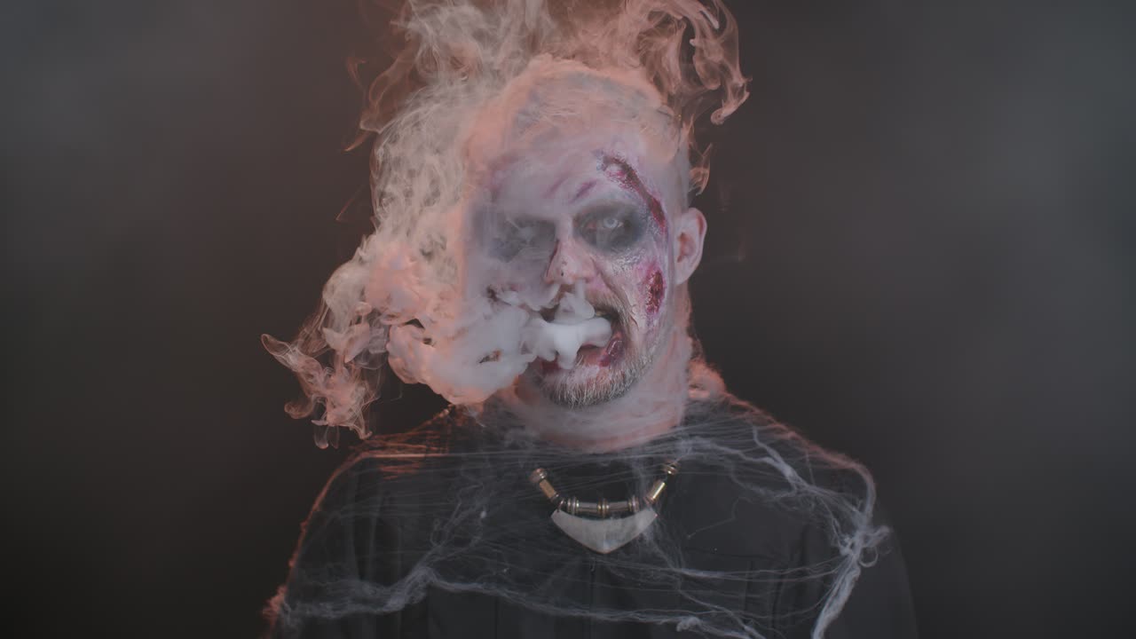Sinister man horrible scary Halloween zombie looking ominous at camera blows smoke from nose, mouth