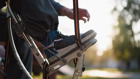 close-up of young man s hands adjusting straps on archery equipment outdoors. he is wearing gray hoodie with soft sunlight illuminating scene, while blurred greenery in background