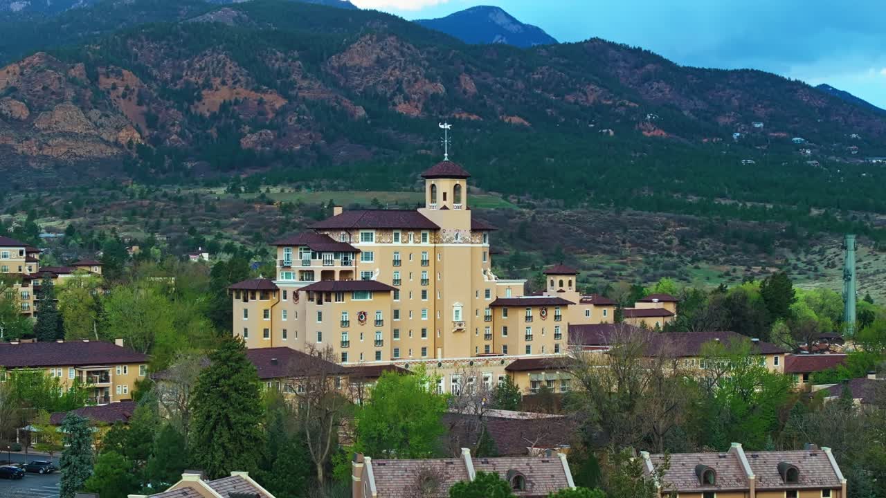 Drone establishing telephoto orbit glides over Broadmoor resort, green trees and lake below under clear sky