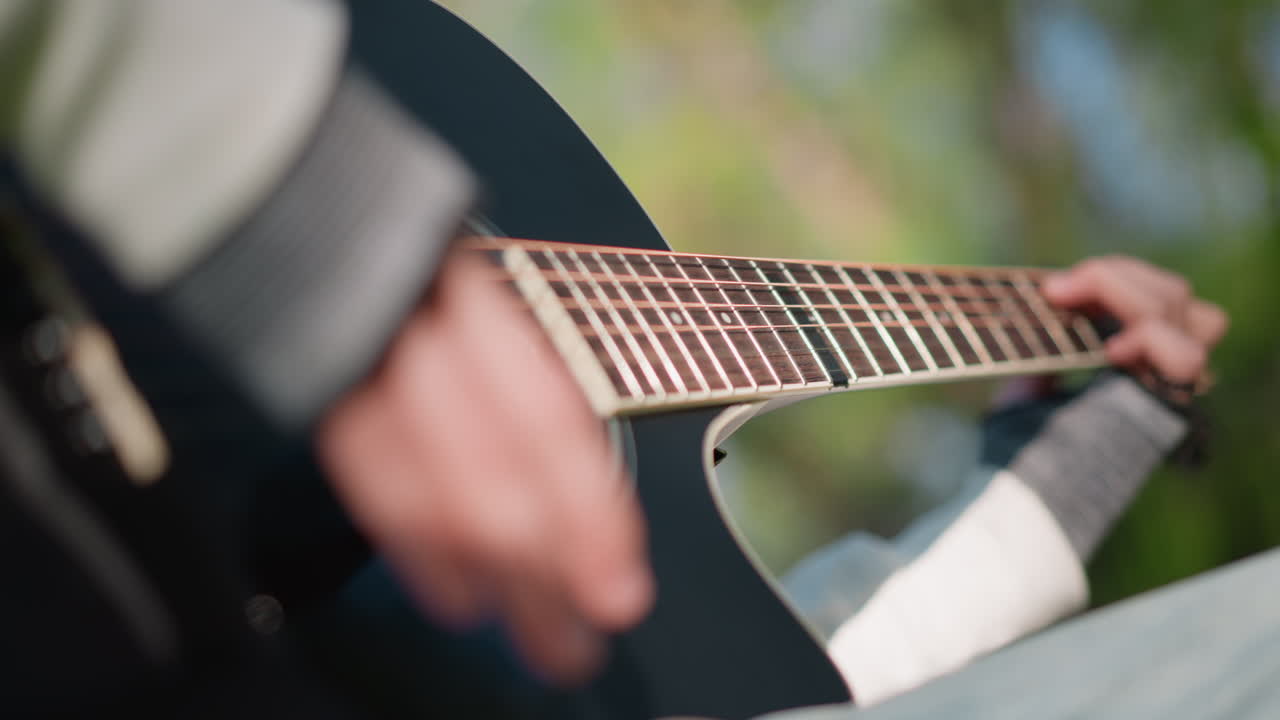 closeup acoustic guitar fretting hands, outdoor daylight with blurred green background, casual sweater, focused fingerpicking and strumming across fretboard, intimate practice session capturing