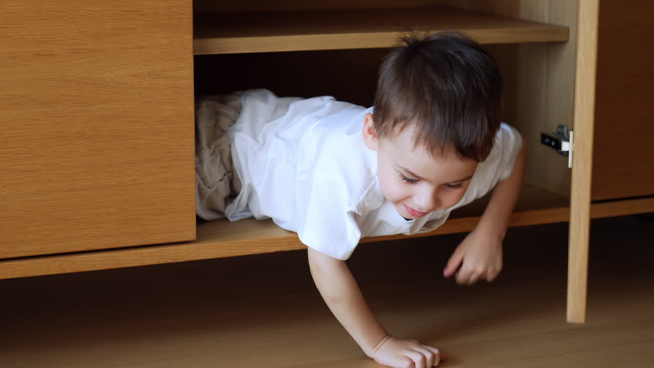 Cute Caucasian toddler, lying inside the nightstand, opens the doors. Dark-haired baby climbs out of chest and approaches a camera smiling