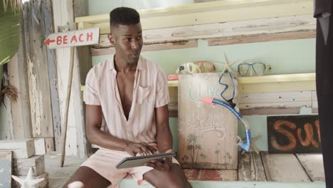 happy african american man using tablet and smiling at surfboard rental beach shack, slow motion