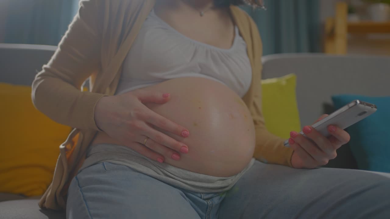 неузнаваемая беременная молодая женщина использует смартфон, сидя на диване дома вблизи.