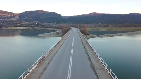 Drone-shot-of-a-girl-riding-towards-the-camera-and-passing-under,-on-a-super-sport-motorcycle-at-a-bridge-over-a-lake-in-Greece