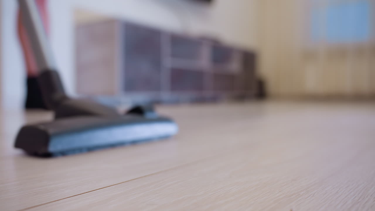 Close up of vacuum cleaner head gliding over light wooden floor with person in black socks standing in blurred background, concept of household chores, cleaning routine and modern home hygiene