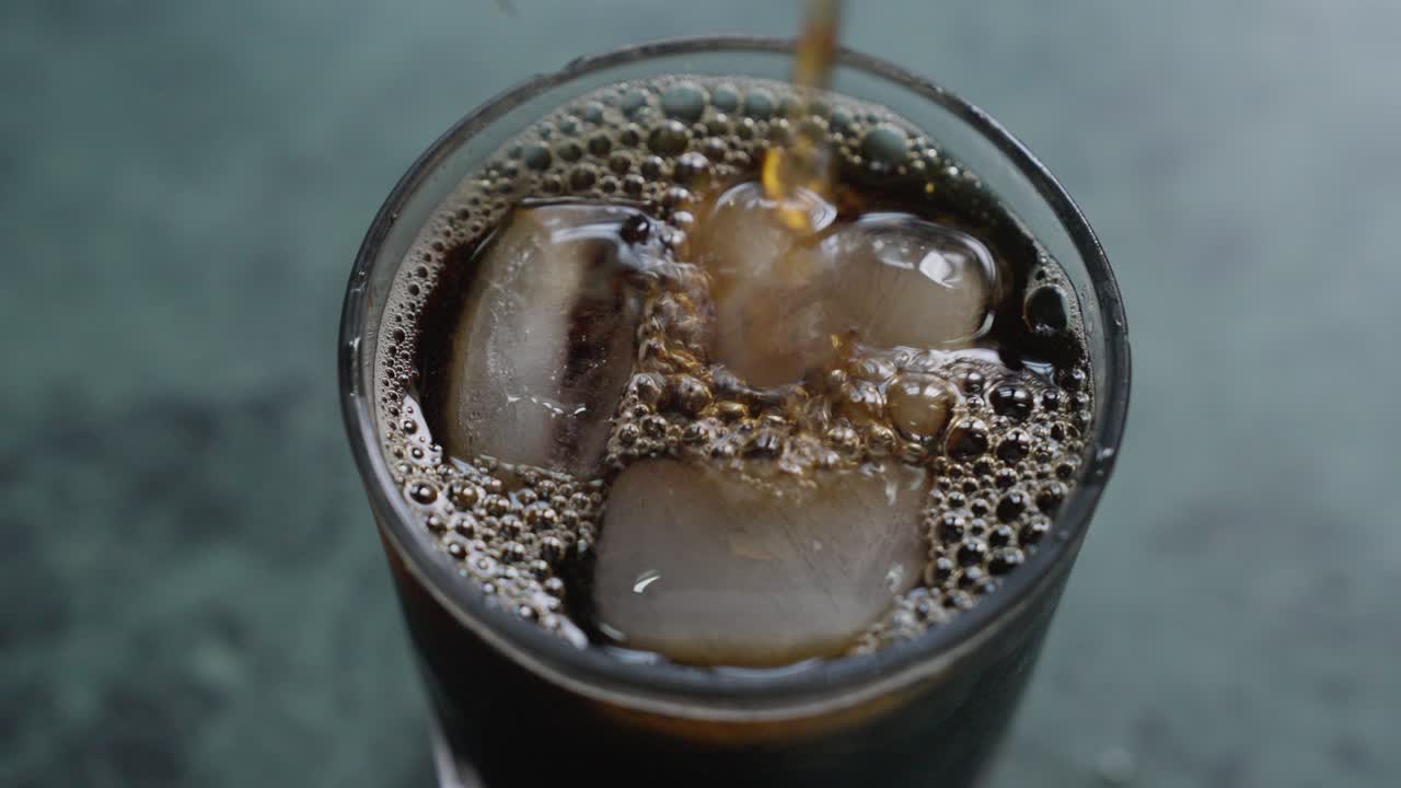 A close-up, top view of pouring iced tea over ice cubes in a transparent glass, with blurred background