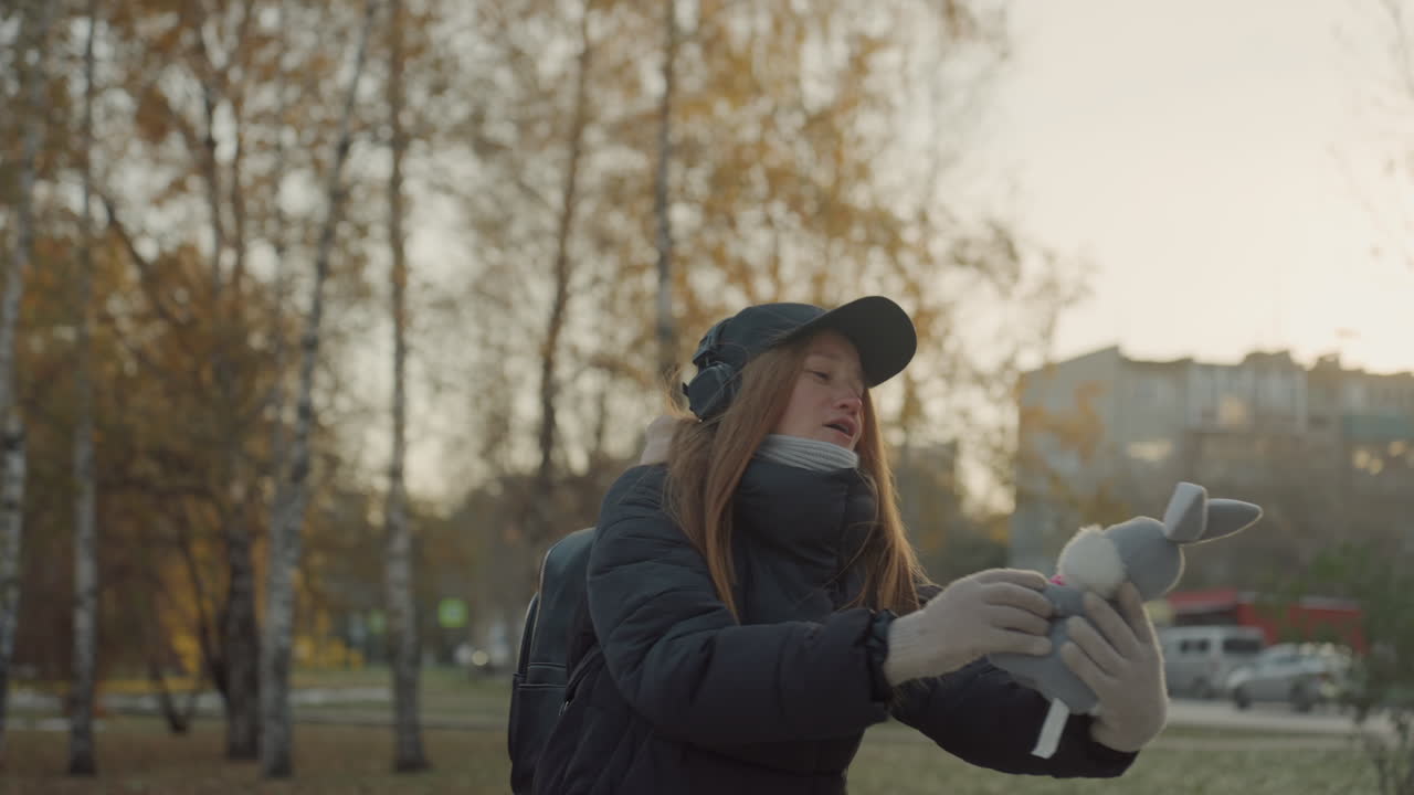 Nainen ottamassa takaisin pehmopupuansa ulkona, nostaen lelun korkealle hämärän valossa iloisella hymyllä, pipo, huivi, hansikkaat ja kuulokkeet yllään, reppu näkyvissä, koivuja ja kaupungin siluetti taustalla.