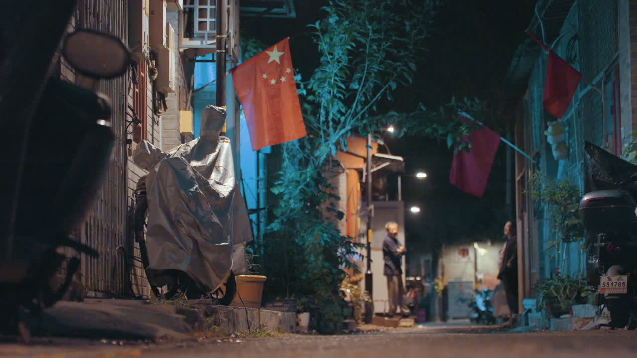 Asılı bayrak ve üzeri örtülü motosiklet bulunan alçak açılı bir ara sokak, ön planda branda ve plastik örtüler, uzakta sıcak lamba ışığının altında durmuş bir kurye, ıslak kaldırım su birikintileri, pürüzlü doku ve neon kontrastı.