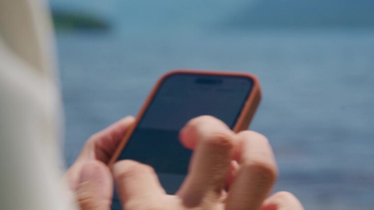 Primo piano di mani che digitano su uno smartphone vicino all'acqua. Le dita toccano lo schermo con concentrazione. Persona rilassata, che si gode il momento al mare. Ideale per social media, viaggi o temi di comunicazione.