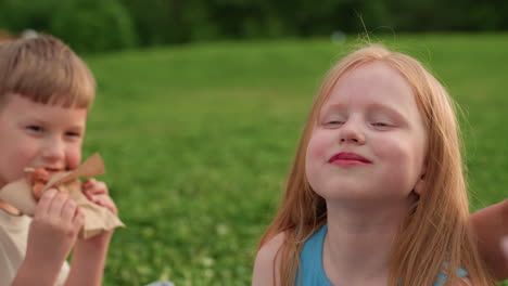 playful girl portrait enjoying picnic with family on green grass, smiling with mouth full of snack, relaxed summer afternoon, closeup showing carefree expression and warm light