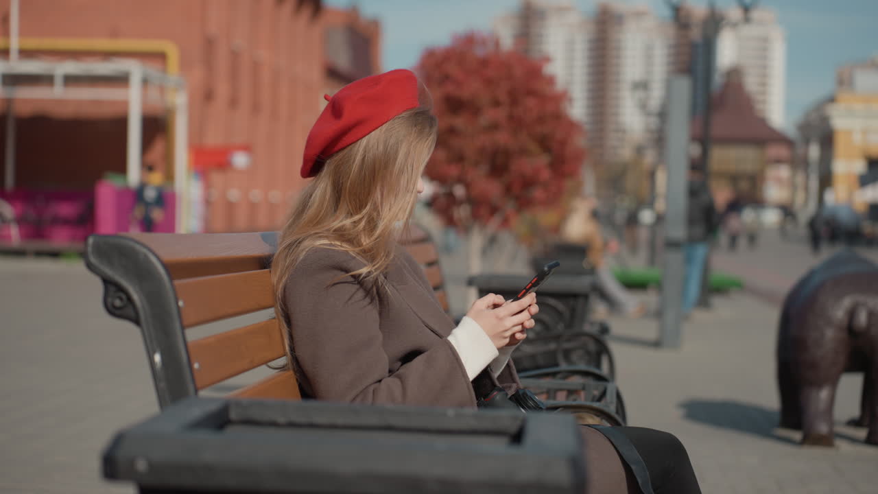 Donna che scrive messaggi all'aperto. Scena autunnale di città con una donna. Strada tranquilla con donna in cappotto. Scena di un parco cittadino isolato con una donna su una panchina. Pomeriggio autunnale con donna che legge messaggi su una panchina del parco in città.