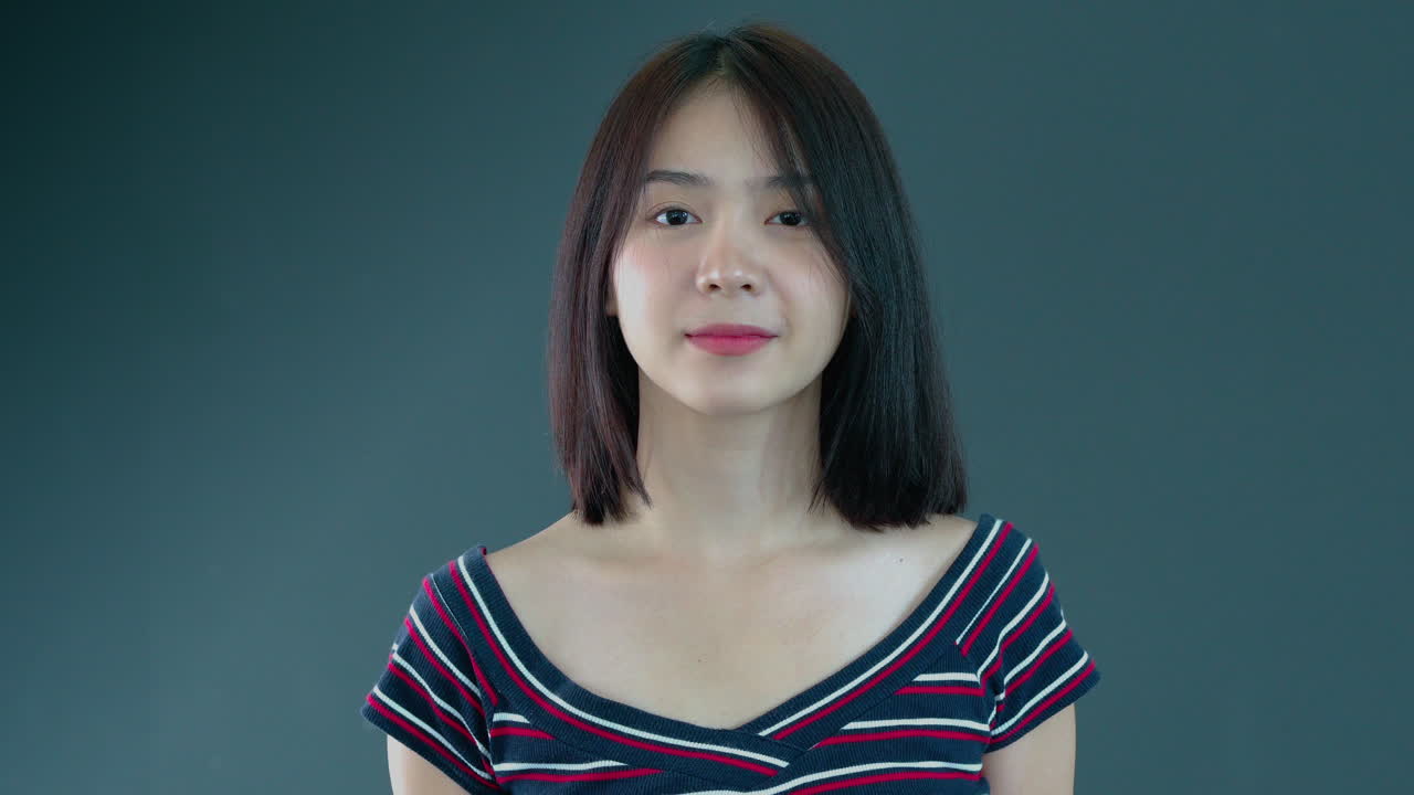 회색 배경에 자신감 있게 웃는 카메라를 바라보는 젊은 아시아 10대 여성의 초상화를 닫습니다