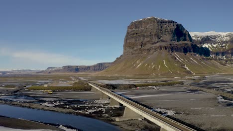 Toma-Aérea-Cinematográfica-Hacia-Atrás-Del-Idílico-Paisaje-Islandés,-Con-El-Delta-Del-Río,-El-Puente-De-Cruce-De-Automóviles-Y-El-Monte-Gigante-Lomagnupur-Como-Telón-De-Fondo-Durante-La-Luz-Del-Sol-En-Islandia