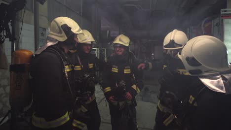 group of five firefighters wearing full equipment in smokey area. relaxed chatting after work well done. fire trucks in the background. name tag translation is fireman and model's last name.