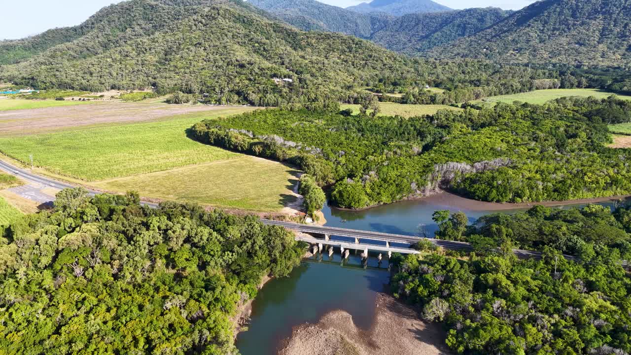 Drone footage captures a vibrant tropical landscape with a river, bridge, and dense greenery under bright sunlight