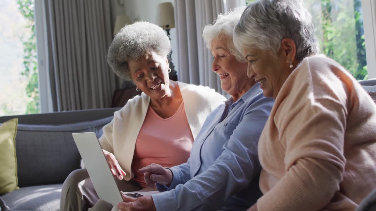Tre donne anziane diverse che usano un laptop insieme sedute sul divano a casa.