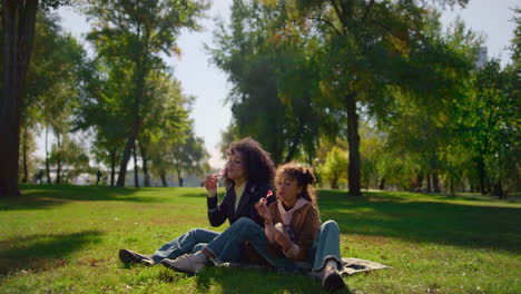 Happy-mother-blowing-soap-bubbles-sitting-on-blanket-in-sunny-park-close-up.