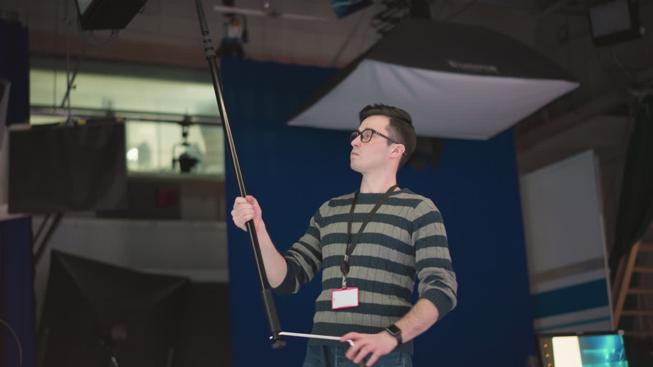 Man holding boom microphone, adjusting it in studio environment with professional lighting, crew, and equipment visible, focusing on sound recording, part of video production process