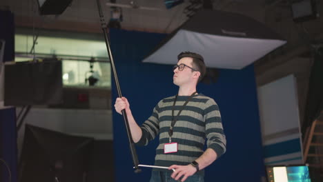 man holding boom microphone, adjusting it in studio environment with professional lighting, crew, and equipment visible, focusing on sound recording, part of video production process