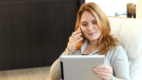 Woman-sitting-on-sofa-using-digital-tablet-while-talking-on-mobile-phone