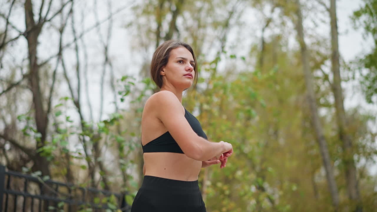 Young lady stretching her body gently outdoors with trees in background, promoting flexibility and wellness in natural environment, focused on maintaining balance and health through exercise
