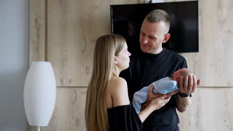 man is holding his newborn son a kissing his wife standing beside. happy new-made parents with their child.