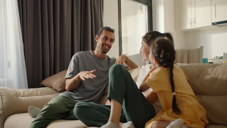 comunicación familiar en el sofá, el padre de una pequeña chica morena en una camiseta gris se comunica con su madre morena mientras está sentado en el sofa y relajándose en una habitación moderna