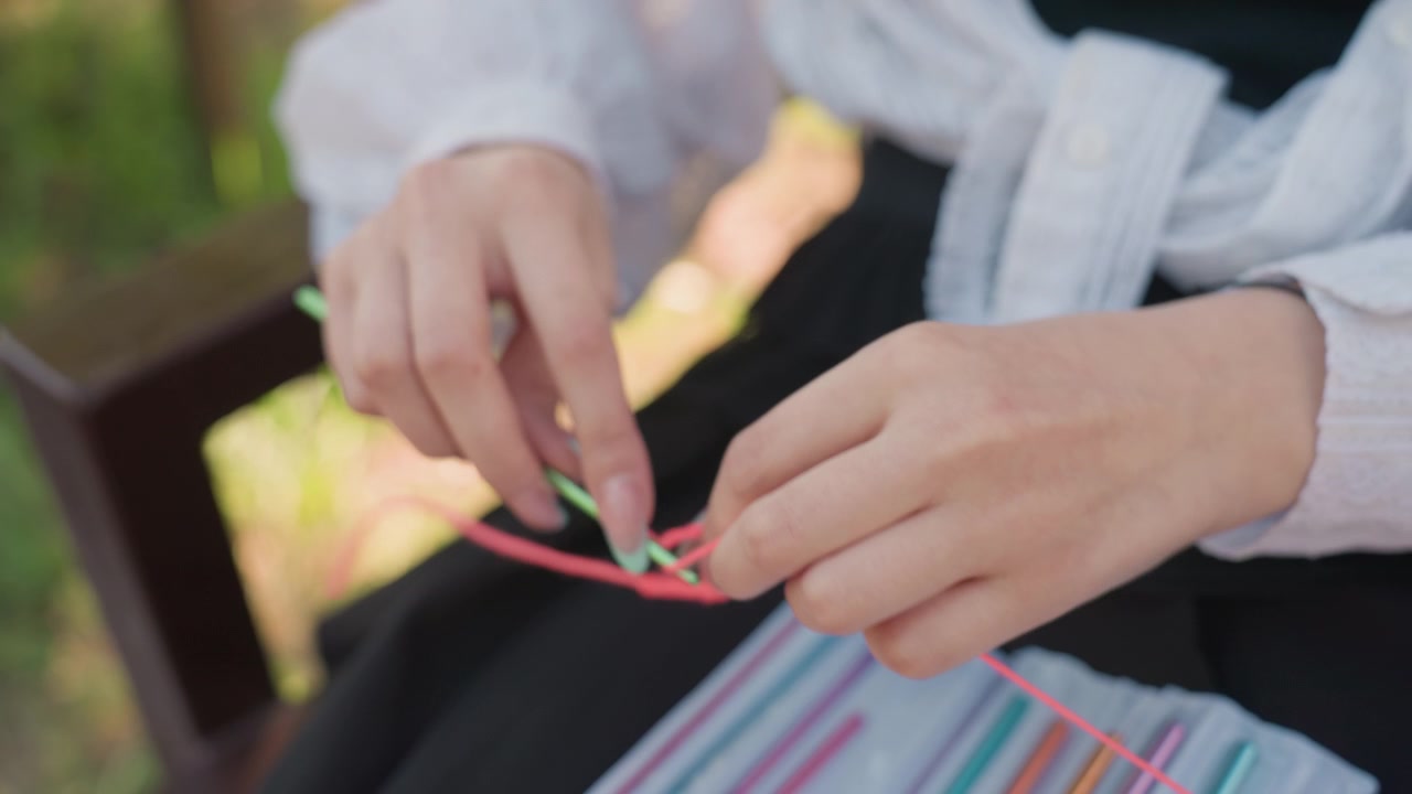 Primo piano di giovane donna che lavora all'uncinetto con filato rosa usando un uncinetto verde, mani attivamente impegnate in un lavoro tessile. Luce naturale soffusa illumina la scena all'aperto, rivelando la mano concentrata