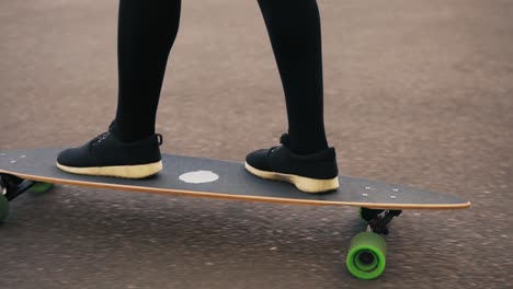 Vista-De-Cerca-De-Las-Piernas-De-Una-Mujer-Con-Zapatillas-Negras-Patinando-En-La-Carretera-De-La-Ciudad.-Piernas-En-El-Monopatín.-Tiro-En-Cámara-Lenta