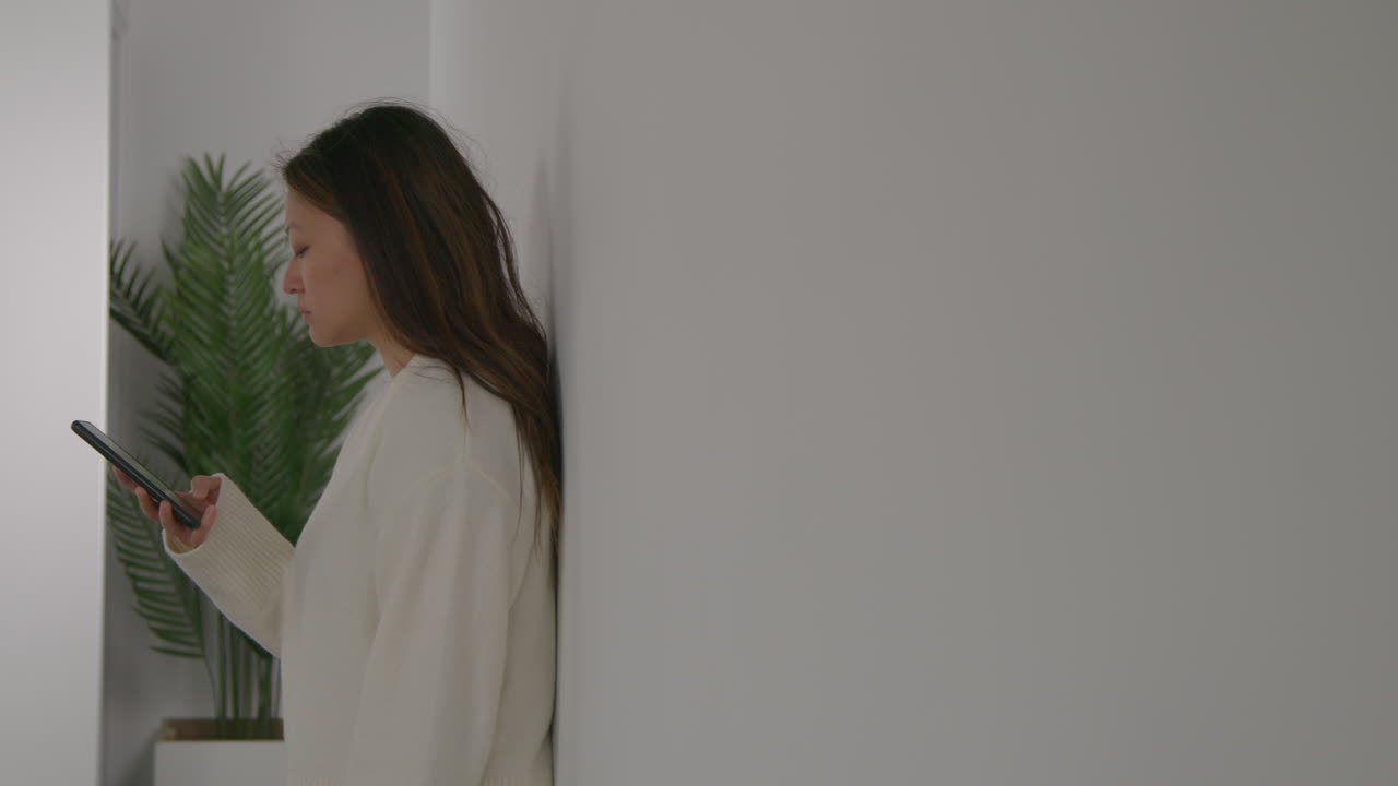 스트레스를 받거나 불안한 여성이 집에서 벽에 기대어 인터넷이나 소셜 미디어 뉴스 메시지 또는 휴대 전화에 반응합니다.