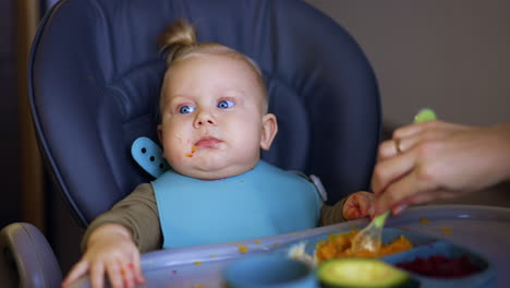 blue-eyed blond infant with food around his mouth sits in high chair. mother gives pureed vegetables to a child.