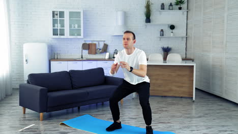 home workout. young man in white t-shirt and black pants doing squats exercises in kitchen interior. sports training indoors. slow motion.