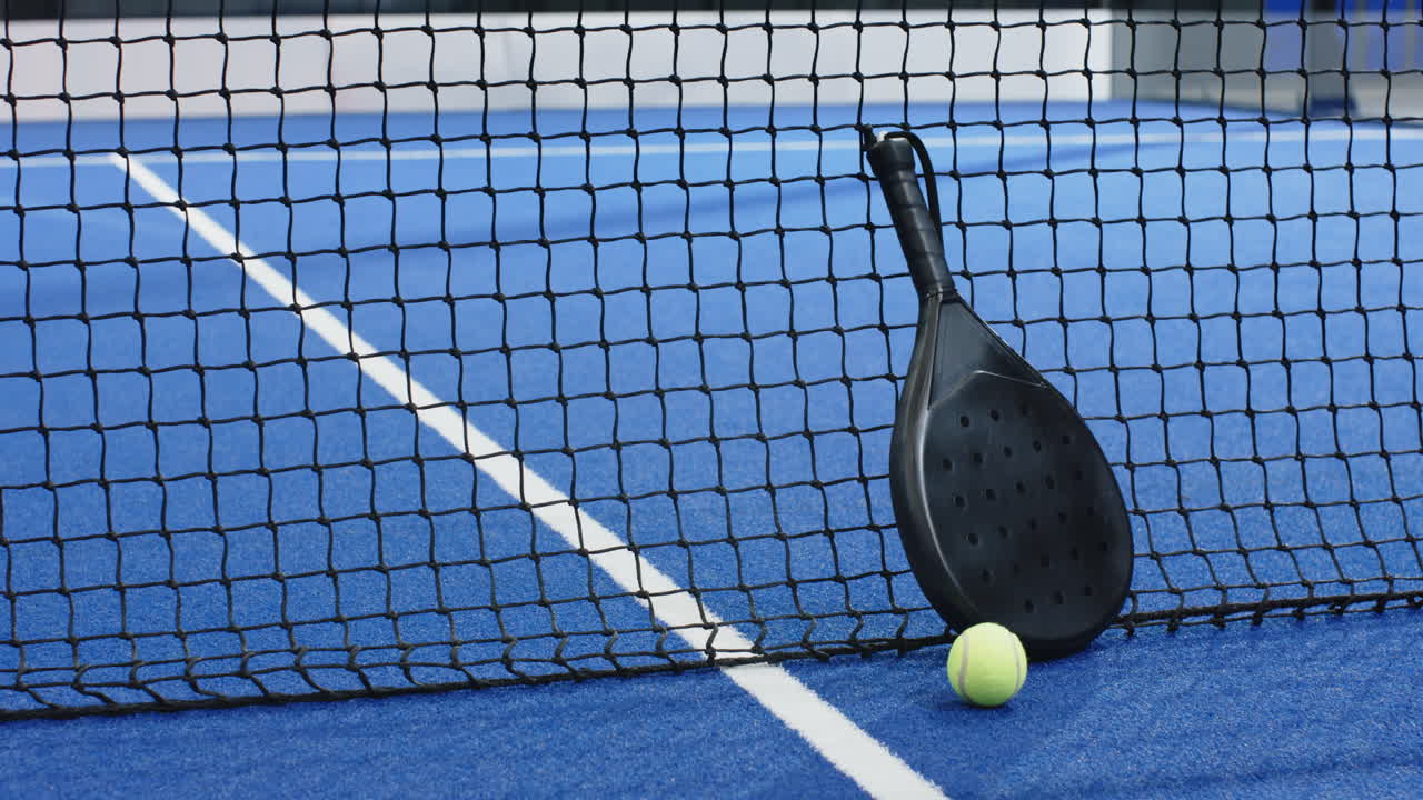 Padel racket and ball resting against net on blue court, copy space