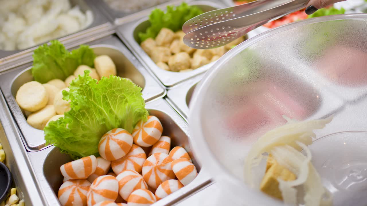 Hand uses tongs to pick seafood and vegetables from buffet trays into a clear bowl