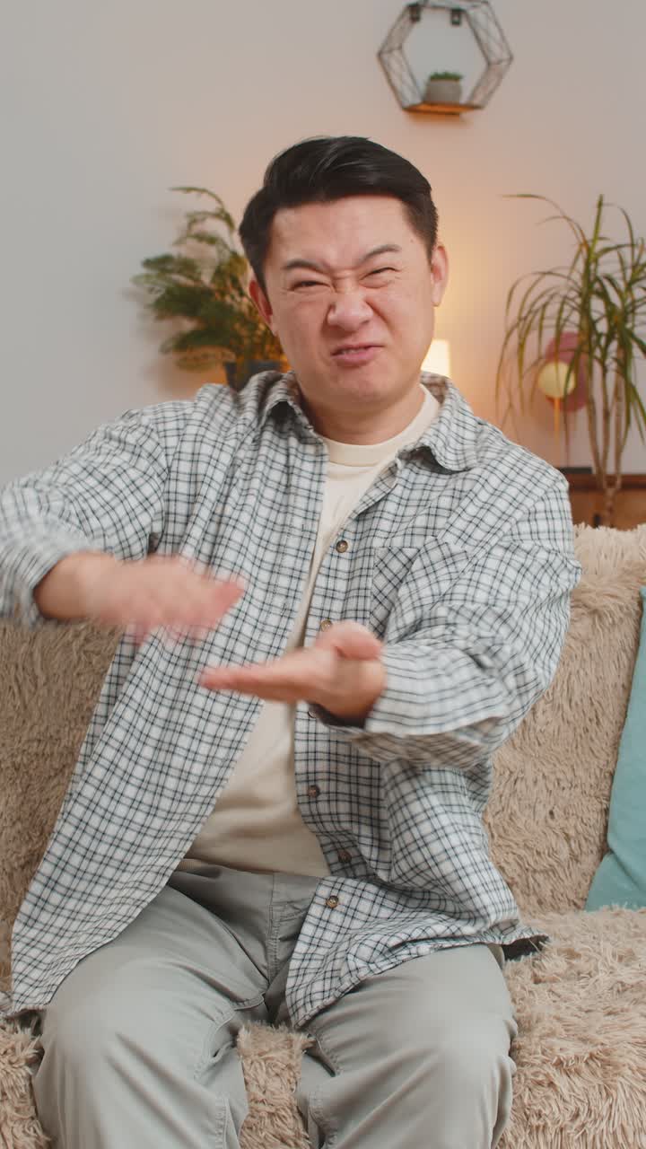 Cheerful rich asian man showing wasting throwing money around hand gesture sitting on sofa at home