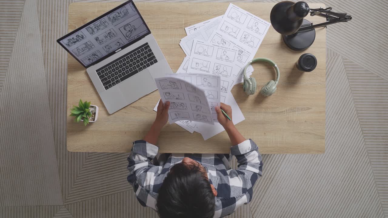 vista superior de artista masculino asiático mirando sus papeles de dibujo eligiendo el mejor guión gráfico para su proyecto en la mesa con una computadora portátil y auriculares en el estudio