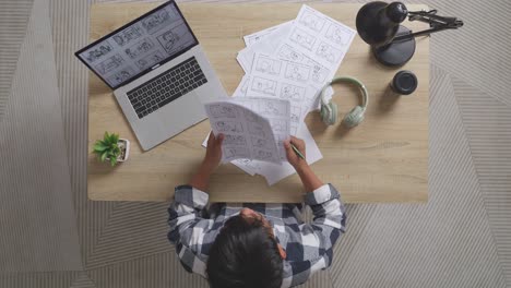 top view of asian male artist looking at his sketching papers choosing the best storyboard for his project on the table with a laptop and headphones in the studio