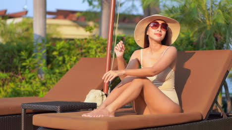 adorable luxury asian lady suntanning on lying on the deck chair at tropical thailand resort wearing beige monokini, sunglasses, and straw hat