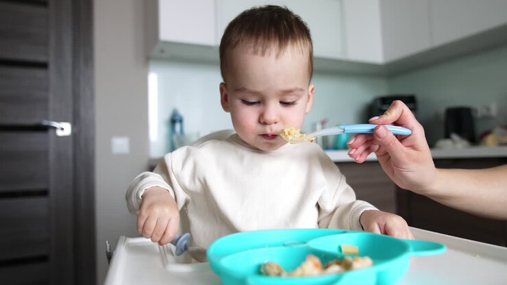 Caucasian baby is fed from a spoon and plate by mom. Kid takes a spoon and tries to pick some food.