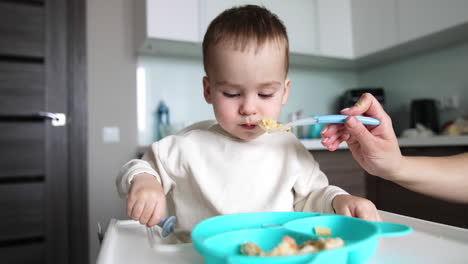 caucasian baby is fed from a spoon and plate by mom. kid takes a spoon and tries to pick some food.