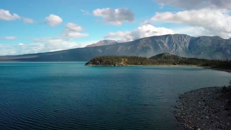 summertime seaside flight above yukon blue turquoise kluane lake shoreline water toward jacquot island and stunning mountain range on bright sunny sky day, canada, overhead aerial approach