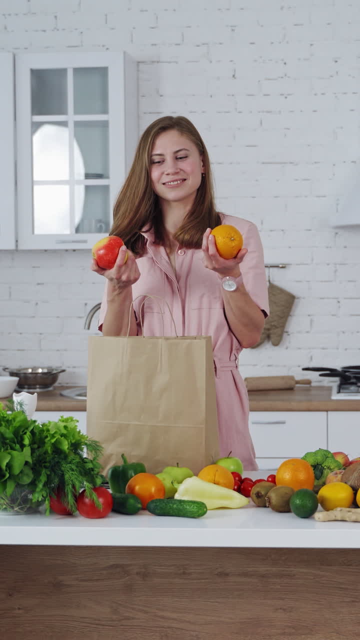 Woman with orange and apple in her hands indoors. Beautiful housewife chooses an apple to eat. Healthy food concept. Vertical video