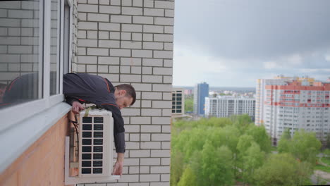 a man installs an external air conditioning unit outside the window of an apartment on a high floor in a big city